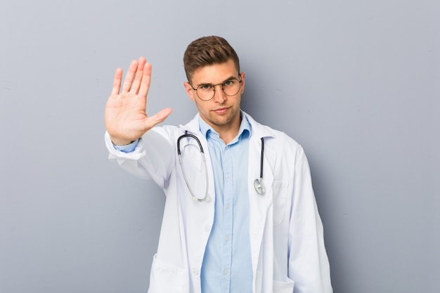 Young blonde doctor man standing with outstretched hand showing stop sign, preventing you.