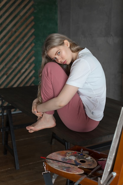 Young blonde female artist sitting on a table near an easel with paints, brush and palette