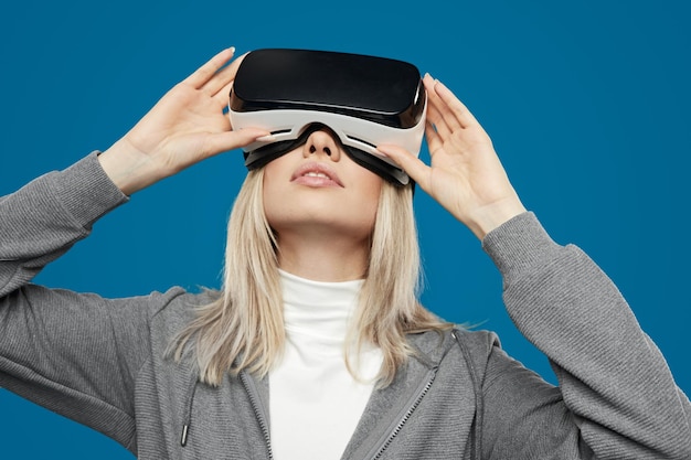 Young blonde female in casual clothes touching VR headset while experiencing virtual world against blue background