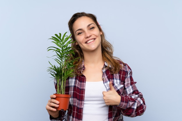 Молодая блондинка садовник девушка держит растение с пальцами вверх, потому что случилось что-то хорошее