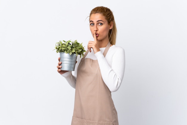 Young blonde gardener woman girl holding a plant over white wall doing silence gesture