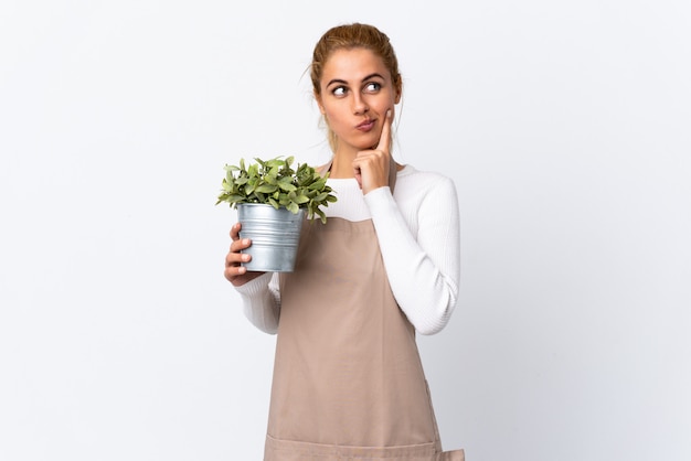 Young blonde gardener woman girl holding a plant over white wall thinking an idea