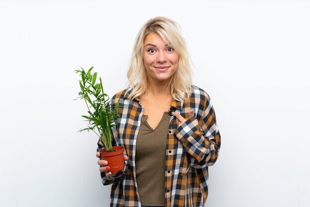 놀라운 표정으로 고립 된 흰 벽 위에 식물을 들고 젊은 금발의 정원사 여자