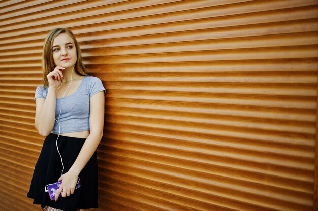 Young blonde girl in black skirt listening music from headphones of mobile phone against orange shutters.