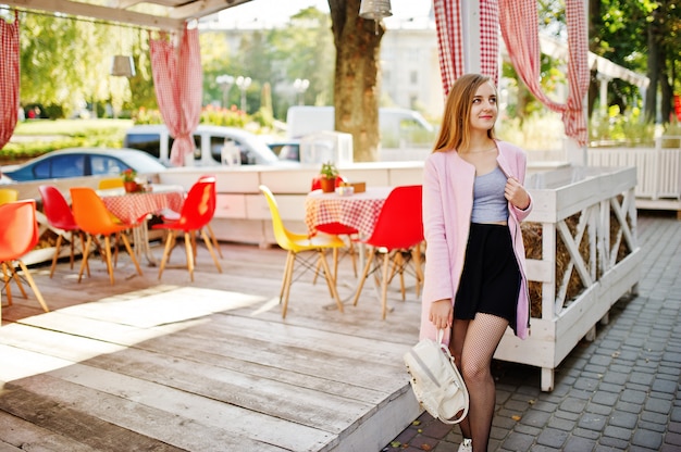 La giovane ragazza bionda in gonna nera e cappotto rosa con lo zaino della donna ha posato contro il ristorante del terrazzo dell'estate all'aperto.