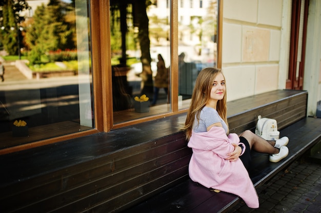 Young blonde girl in black skirt and pink coat with woman backpack sitting on bench at city.