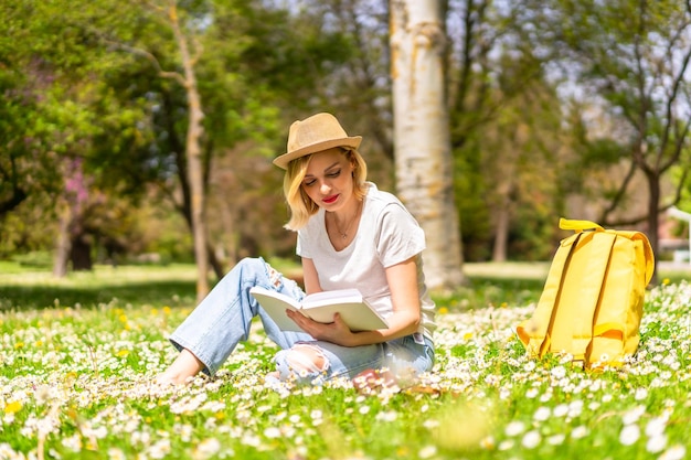 A young blonde girl in a hat reading a book in spring in a park\
in the city vacations next to nature and next to daisies