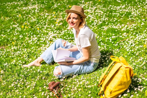 A young blonde girl in a hat writing poems in a notebook in
spring in a park in the city nature sitting on the grass next to
daisies