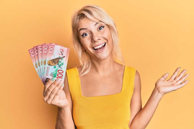 Молодая блондинка с банкнотой в 100 новозеландских долларов празднует достижение со счастливой улыбкой и выражением лица победителя с поднятой рукой