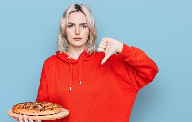 Young blonde girl holding italian pizza with angry face, negative sign showing dislike with thumbs down, rejection concept