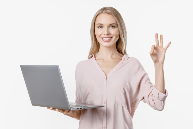 Young blonde girl holding laptop computer and showing ok gesture isolated over white background