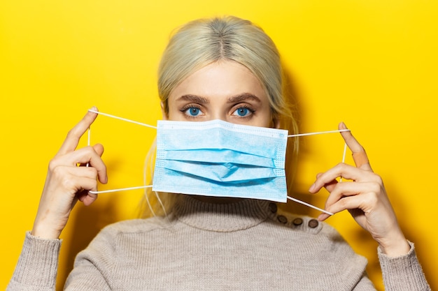 Young blonde girl holding a medical flu mask on face, on yellow wall