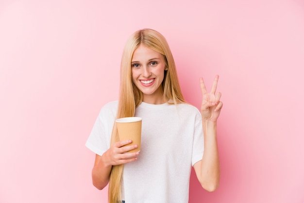 Молодая белокурая девушка держа на вынос кофе показывая знак победы и широко усмехаясь.