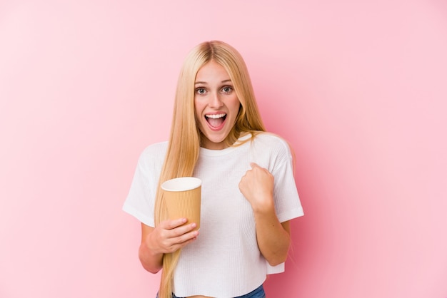 Young blonde girl holding a takeaway coffee surprised pointing at himself, smiling broadly.