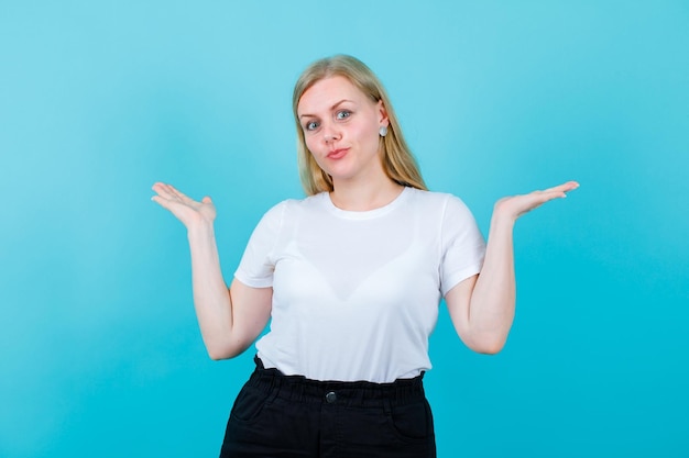 Young blonde girl is looking at camera by opening wide her hands on blue background