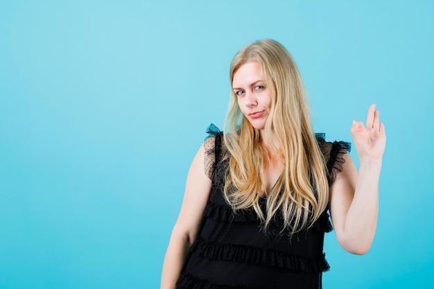 Photo young blonde girl is looking at camera by showing okay gesture on blue background