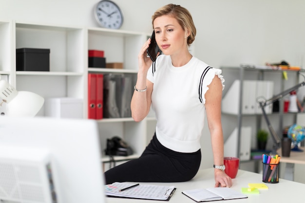 A young blonde girl is sitting at the desk in the office and talking on the phone.