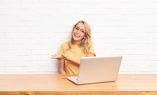 young blonde girl at laptop smiling cheerfully and showing something with the hand