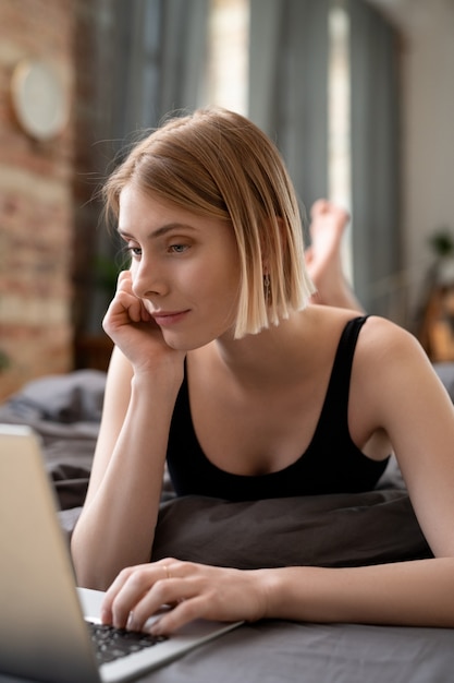 Giovane ragazza bionda sdraiata sul letto e usa il laptop nel suo lavoro nella stanza