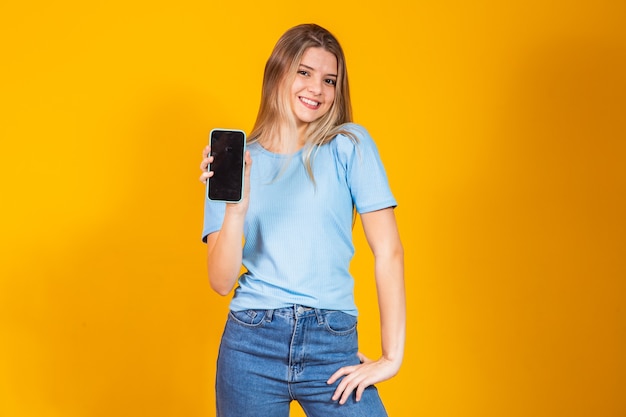 Young blonde girl showing blank smartphone screen on yellow background.