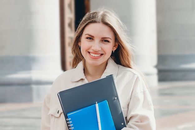 Young blonde girl student smiling against university. Cute girl student holds folders and notebooks in hands. Learning, education concept