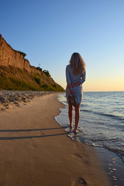 A young blonde girl walks along the beach barefoot and carries shoes in her hand Back view