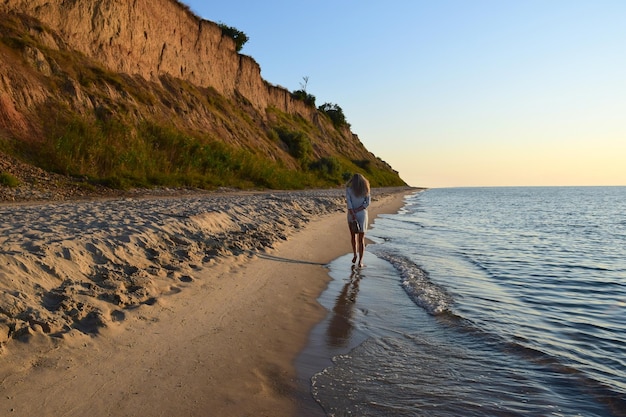 A young blonde girl walks along the beach barefoot and carries shoes in her hand Back view