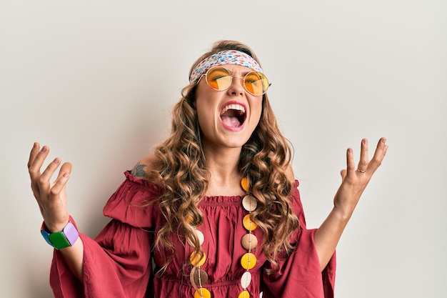 Young blonde girl wearing bohemian and hippie style crazy and mad shouting and yelling with aggressive expression and arms raised frustration concept