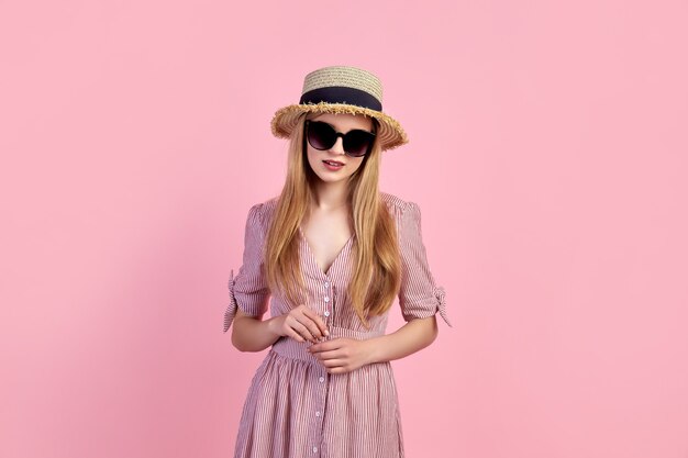 Photo young blonde girl wearing dress, straw hat, isolated over pink background.