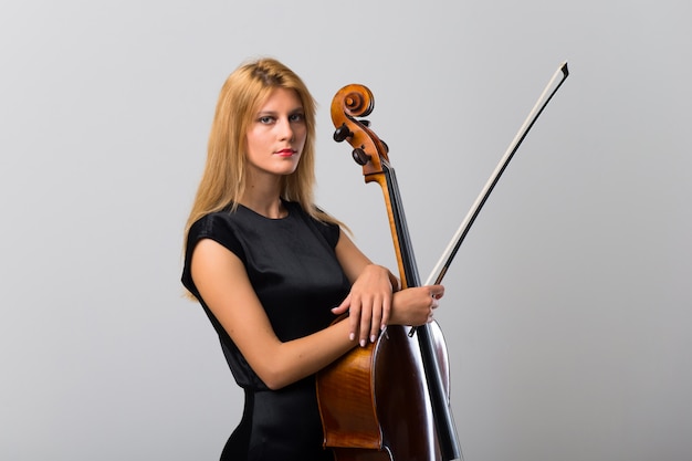 Young blonde girl with her cello posing on white wall