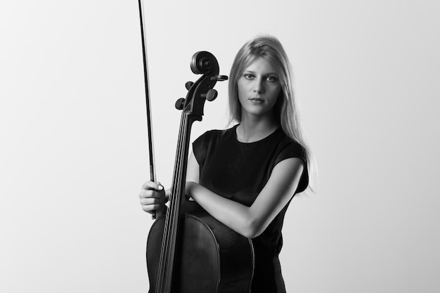 Young blonde girl with her cello posing on white wall