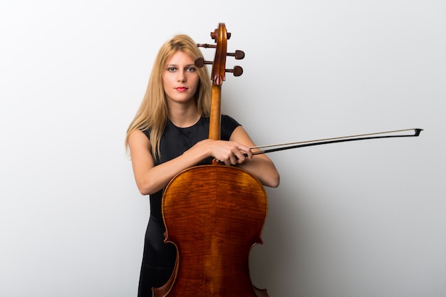 Young blonde girl with her cello posing on white wall