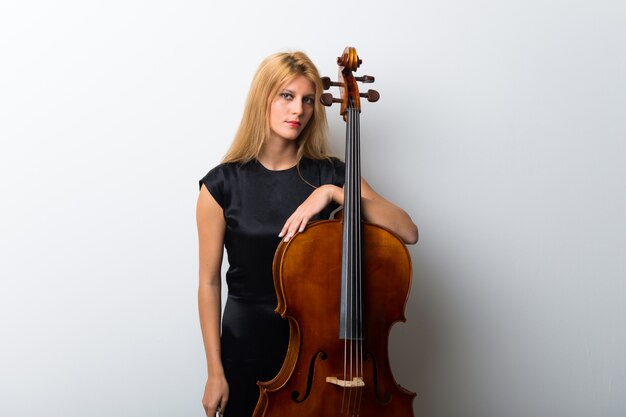 Young blonde girl with her cello posing on white wall