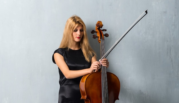 Young blonde girl with her cello on textured wall background