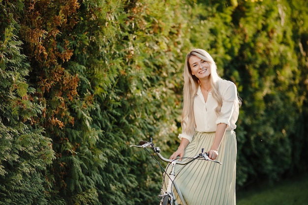 Young blonde girl with long hair standing near vintage white bicycle.