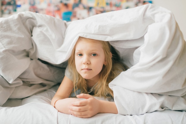 Young  blonde girl with sad face lying on bed under white blanket and look at camera. Little female child with pensive face indoor lifestyle portrait.