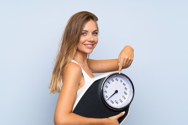 Young blonde girl with weighing machine over isolated blue white