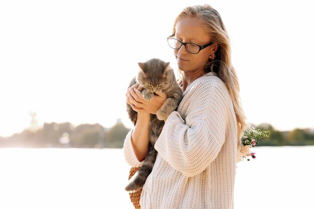 Young blonde girl woman with glasses holds a cat breed Scottish Straight Walk with pet near the lake river at sunset on the beach in Spring