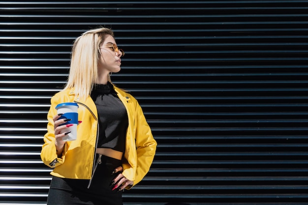 young blonde hispanic latin girl dressed in black and yellow posing with a reusable coffee cup