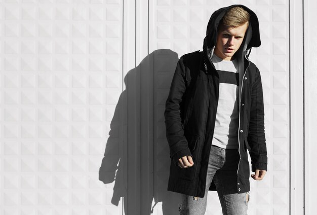 Young blonde man in autumn clothes standing near white wall.