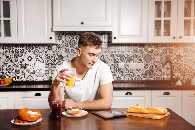 Young blonde man drinking juice and reading news on the tablet\
while having breakfast