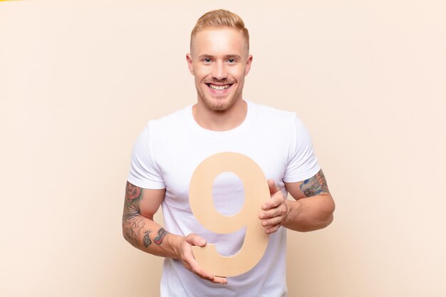 Young blonde man excited, happy, joyful, holding a number 9.