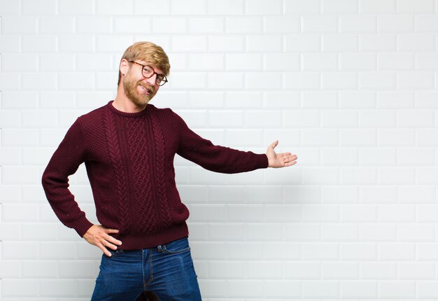 Young blonde man feeling happy and cheerful, smiling and welcoming you, inviting you in with a friendly gesture agaist vintage tiles wall