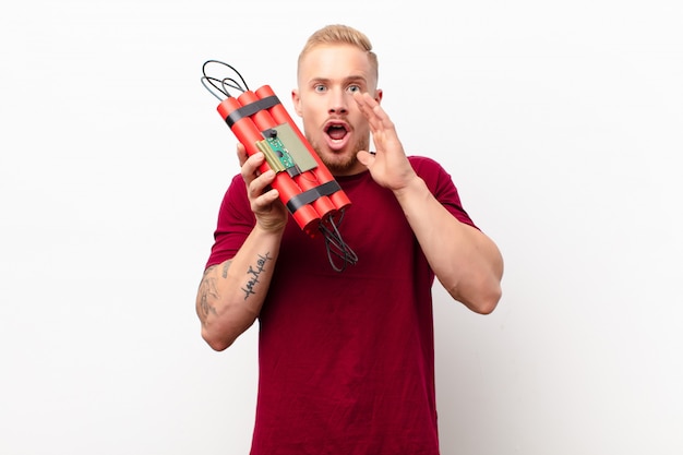 Young blonde man feeling happy, excited and positive, giving a big shout out with hands next to mouth, calling out against white wall holding a dynamite explosive