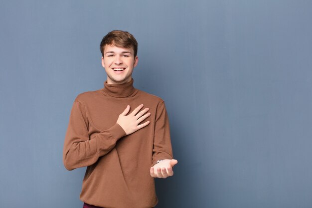 Young blonde man feeling happy and in love, smiling with one hand next to heart and the other stretched up front isolated on flat wall