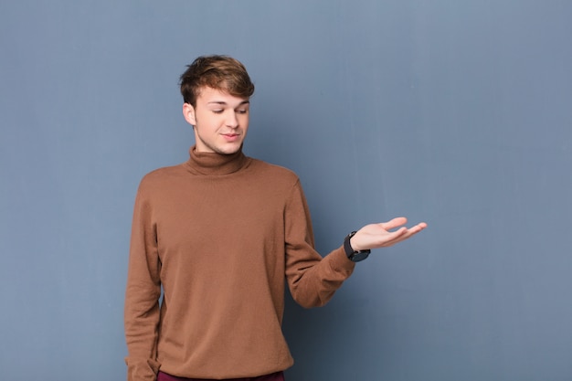 Young blonde man feeling happy and smiling casually, looking to an object or concept held on the hand on the side isolated against flat wall