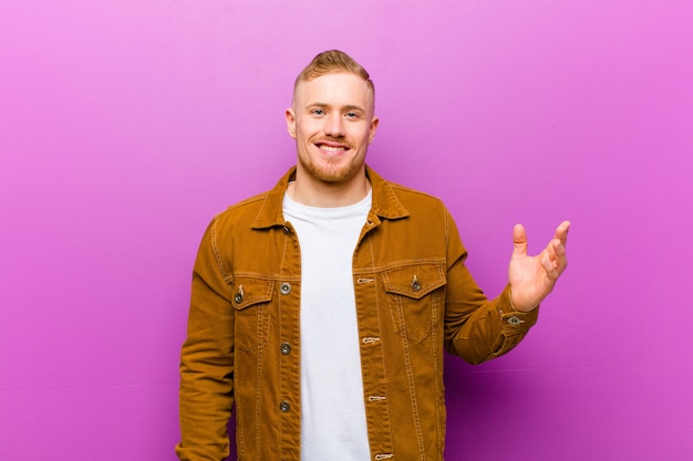 Young blonde man feeling happy, surprised and cheerful, smiling with positive attitude, realizing a solution or idea against purple wall