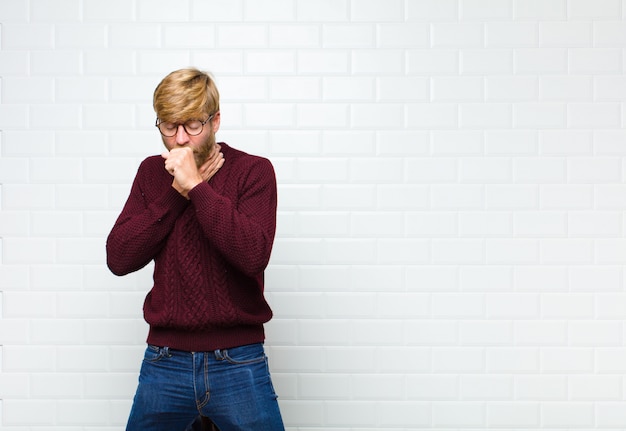 Young blonde man feeling ill with a sore throat and flu symptoms, coughing with mouth covered agaist vintage tiles wall