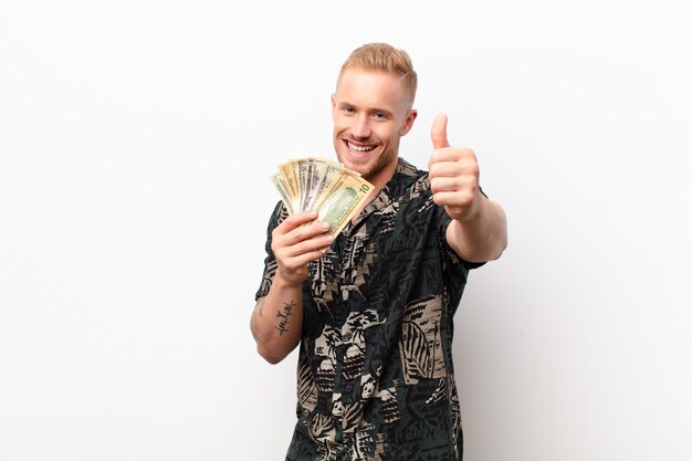 Young blonde man feeling proud, carefree, confident and happy, smiling positively with thumbs up over white wall with banknotes