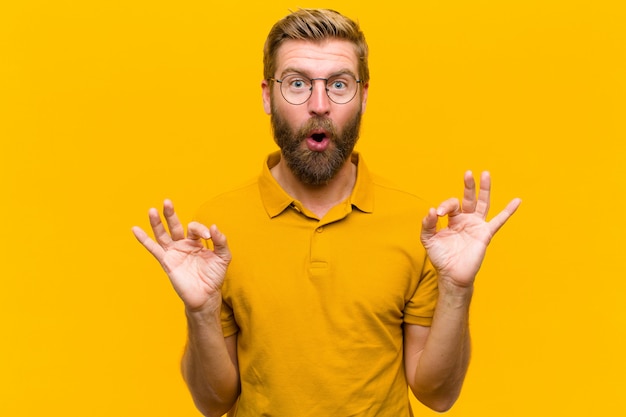 Young blonde man feeling shocked, amazed and surprised, showing approval making okay sign with both hands  orange wall
